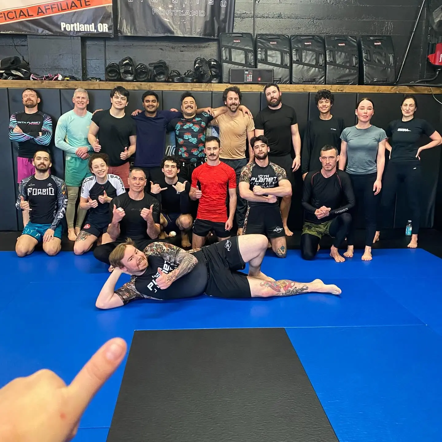 A full weeknight class at 10th Planet Portland lined up on the mats, Thai pads and shin guards stacked on the shelf behind them