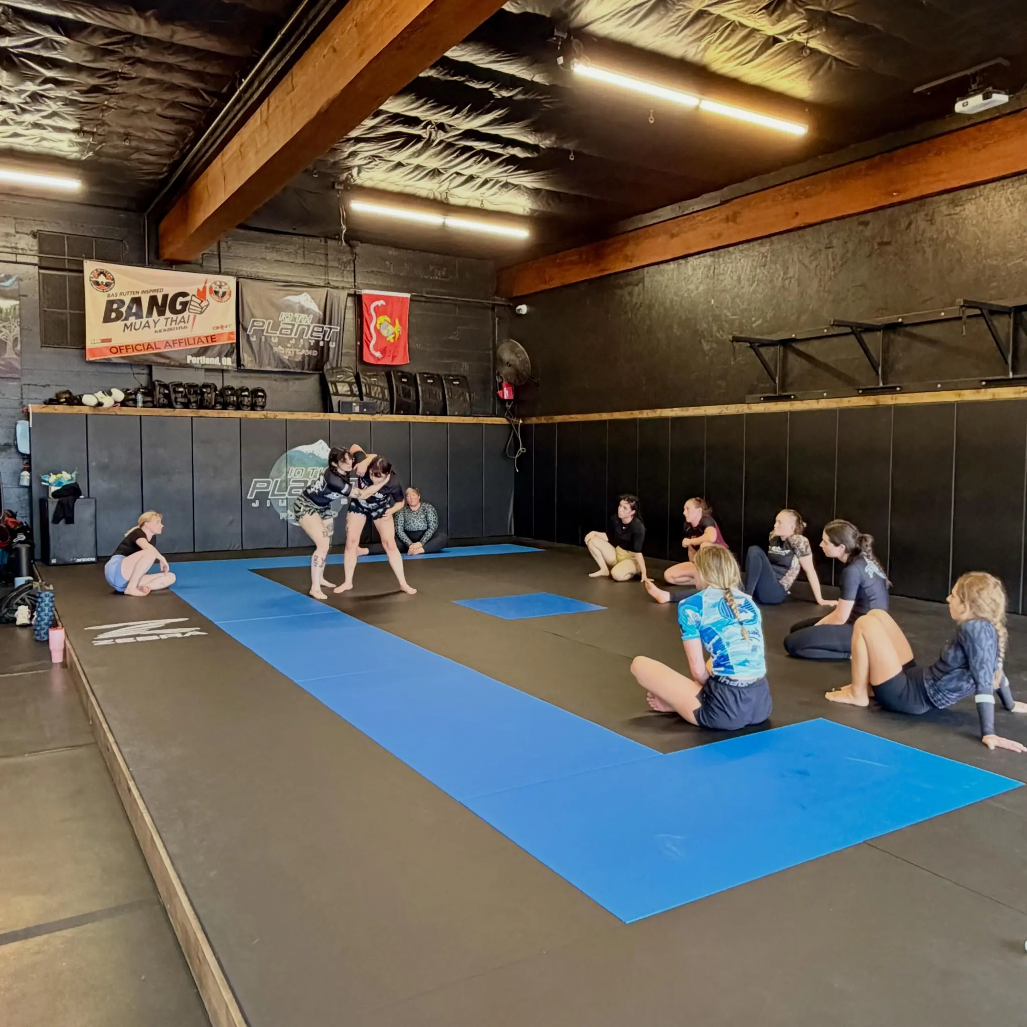 Wide interior view of the 10th Planet Portland mat room, exposed wooden roof beams above a full floor of mats with a class in session