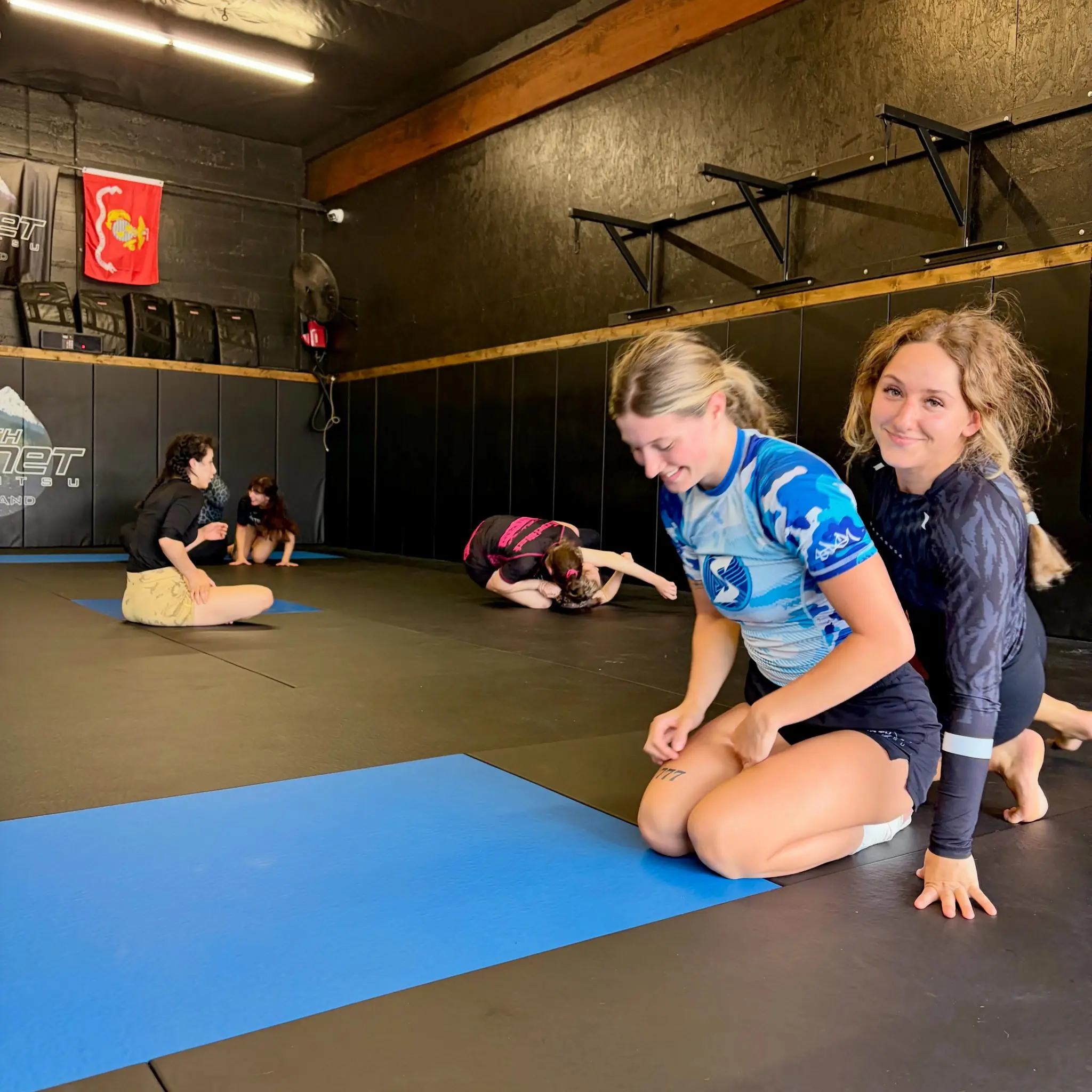 Two women kneeling on the mats smiling during a women's jiu jitsu session while others drill behind them