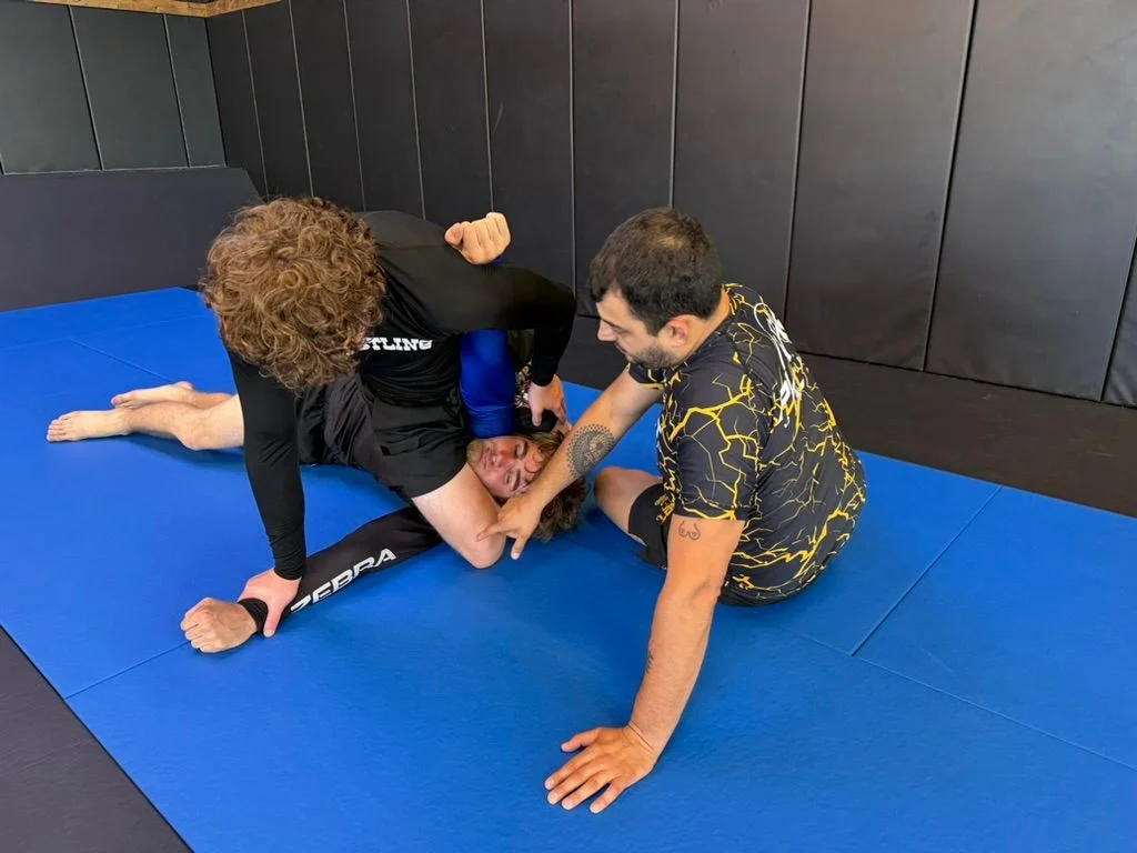A coach at 10th Planet Portland working hands-on with two adult students on a position, kneeling beside them on the blue mats