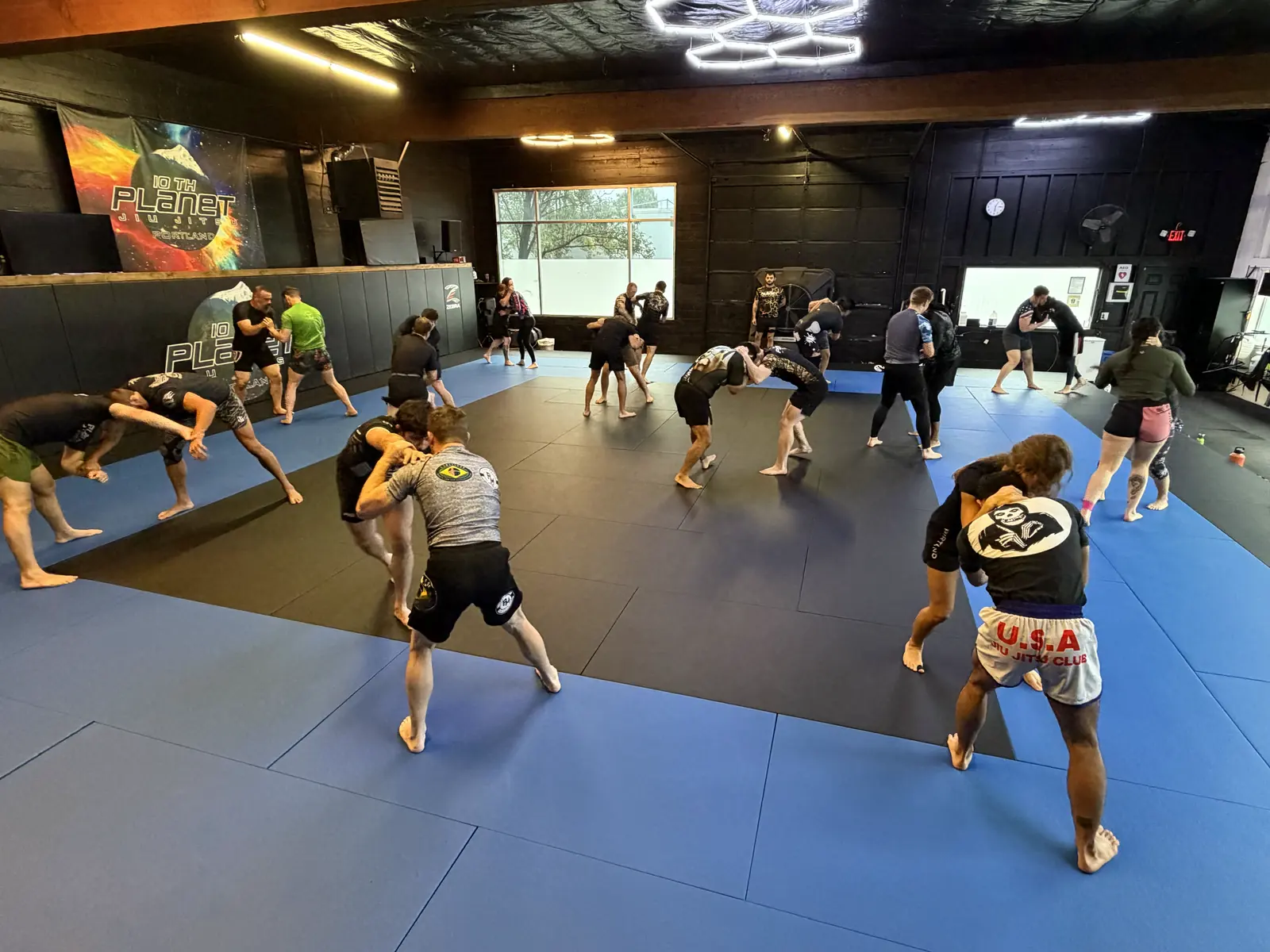 A full adult class at 10th Planet Portland working in pairs across the main mat room, the 10th Planet banner on the padded wall behind them