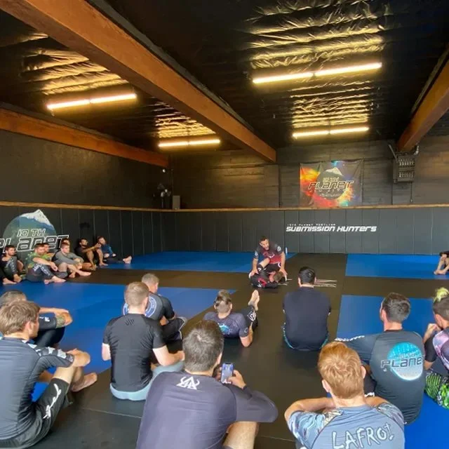 Corner view of the 10th Planet Portland mat room under exposed beams and strip lighting, a full class seated for instruction