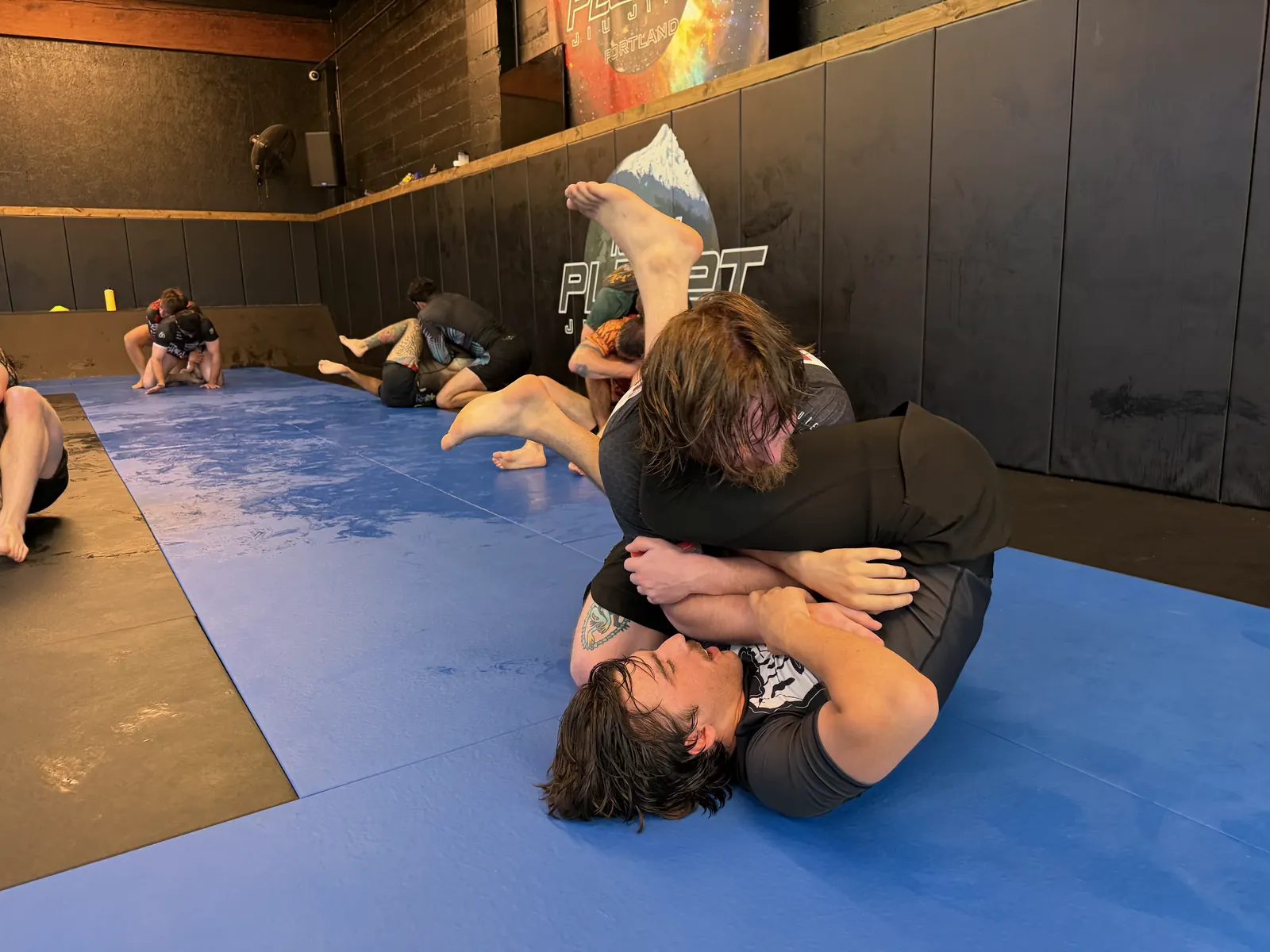 Two adults rolling no-gi at 10th Planet Portland, one working from guard with the 10th Planet wall banner and other pairs training behind them